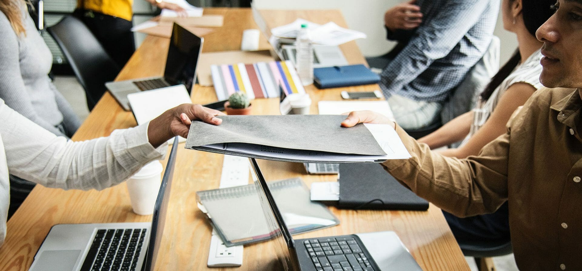 Eine Blended-Learning-Gruppe sitzt an einem Tisch mit Laptops und Dokumenten. Im Vordergrund reicht eine Person ihrem Gegenüber eine Dokumentenmappe.