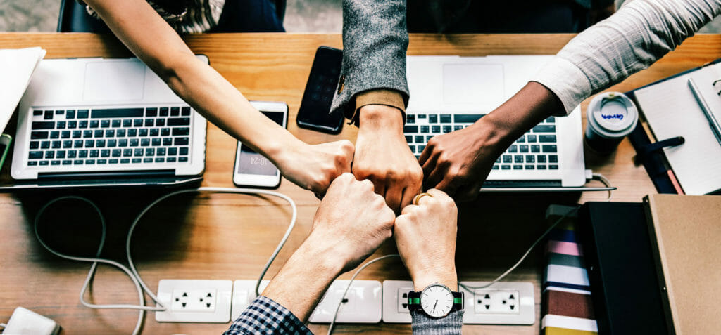 Vijf mensen die rondom een tafel zitten en met laptops aan e-learning deelnemen, ze geven elkaar een fist bump.