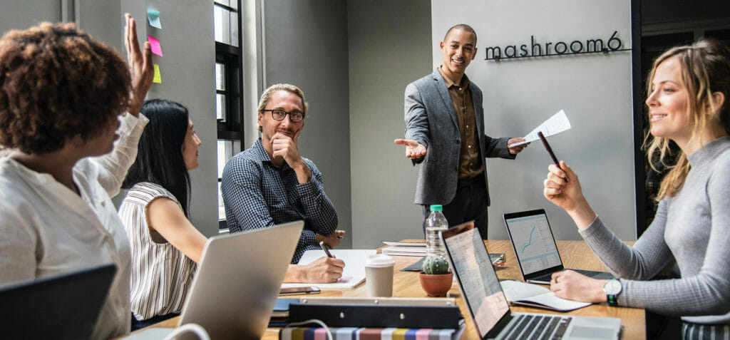 Lab-Rotationsmodell mit 4 Personen an einem Tisch mit Laptops. Eine Frau meldet sich zu Wort. Ein Mann steht vor dem Tisch und macht eine auffordernde Geste.