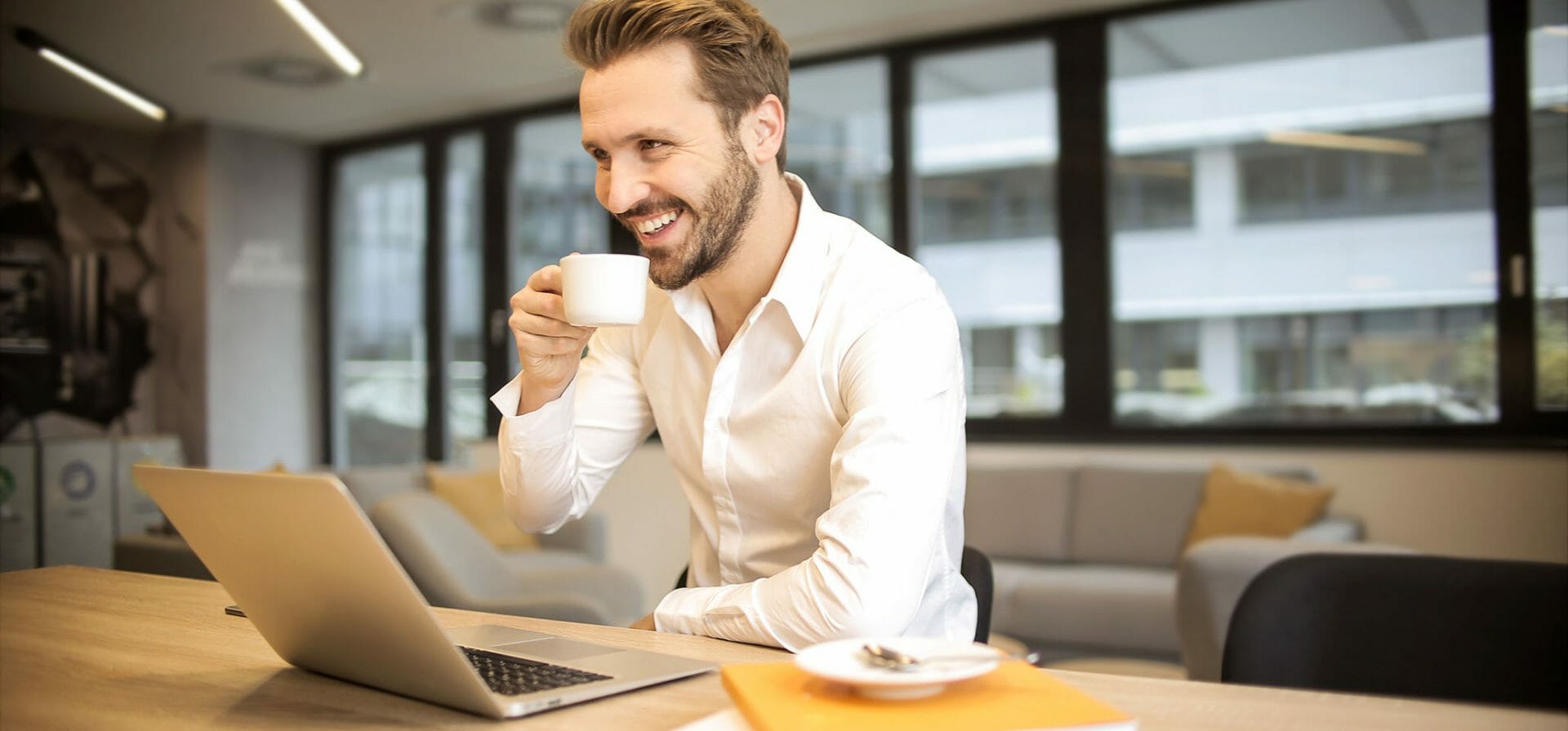 Ein Mann sitzt mit einer Tasse Kaffee vor einem Laptop, rechts daneben eine Untertasse und eine Dokumentenmappe.