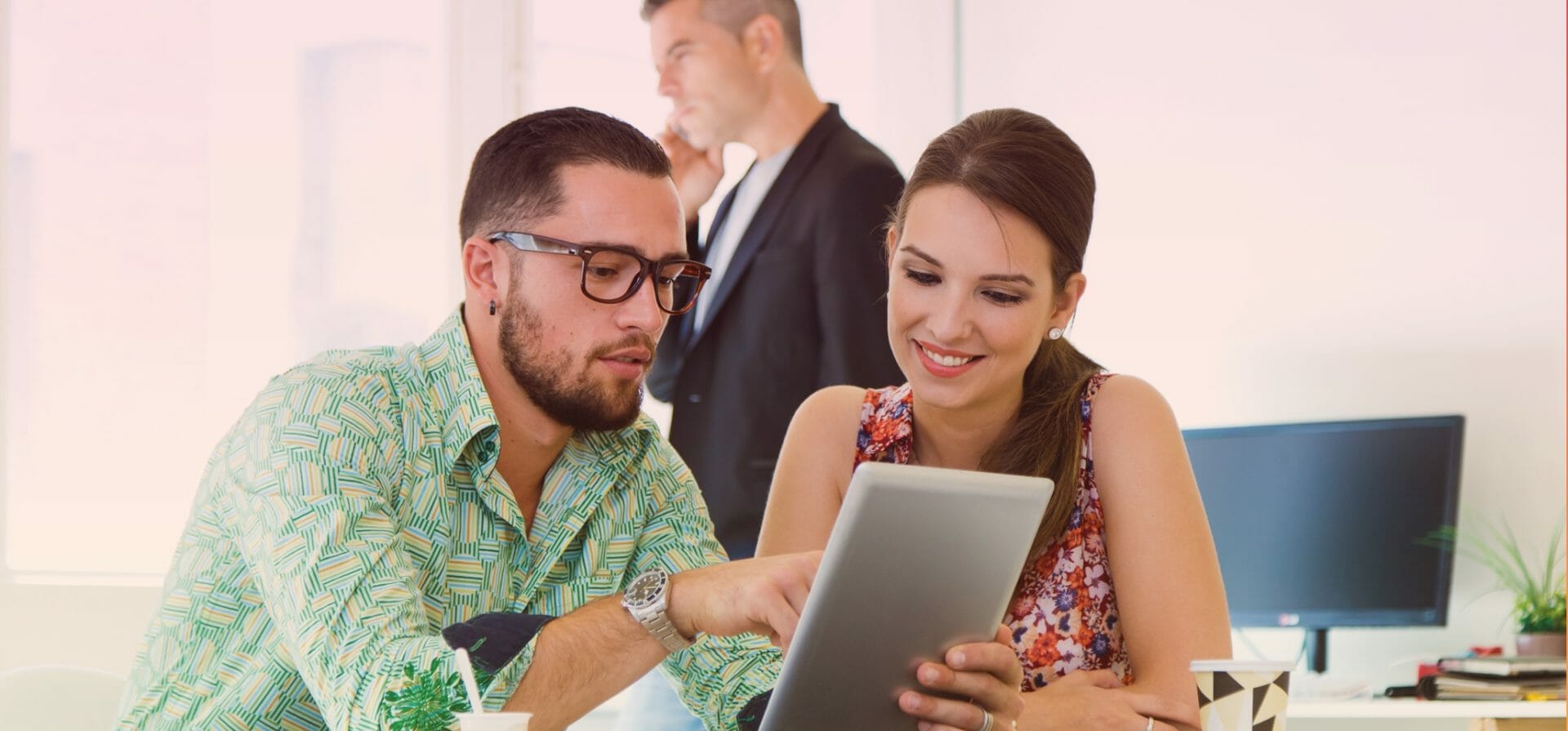 Dos personas jóvenes y sonrientes comparten conocimientos mientras miran la misma tableta electrónica en una oficina.