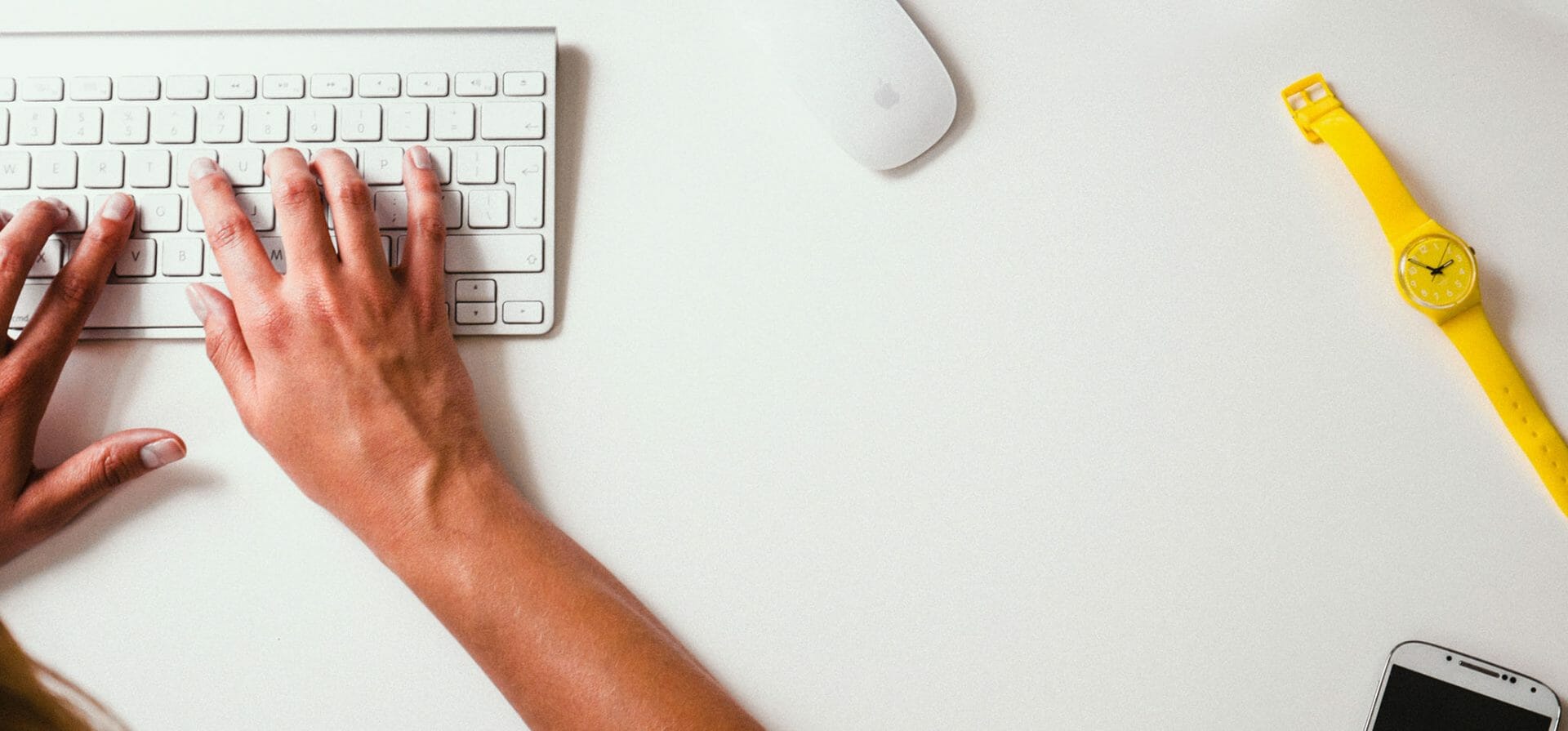 Una persona escribe en un teclado junto a un ratón, un reloj de pulsera amarillo y un teléfono móvil.