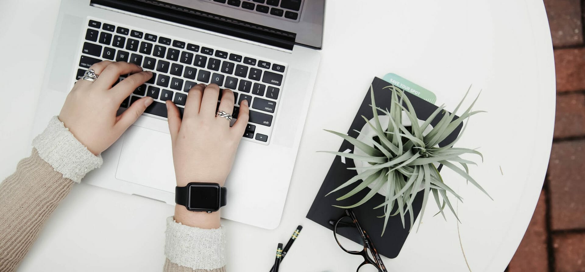 Een persoon met een smartwatch en ringen, werkt aan een LMS op een witte tafel met een laptop, een plant, een bril en twee pennen.