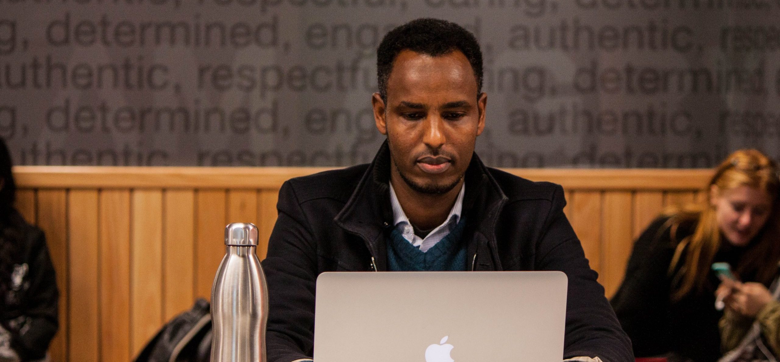 Ein Mann sitzt an einem Tisch vor einem Macbook. Im Hintergrund sind links und rechts 2 sitzende Personen zu sehen.