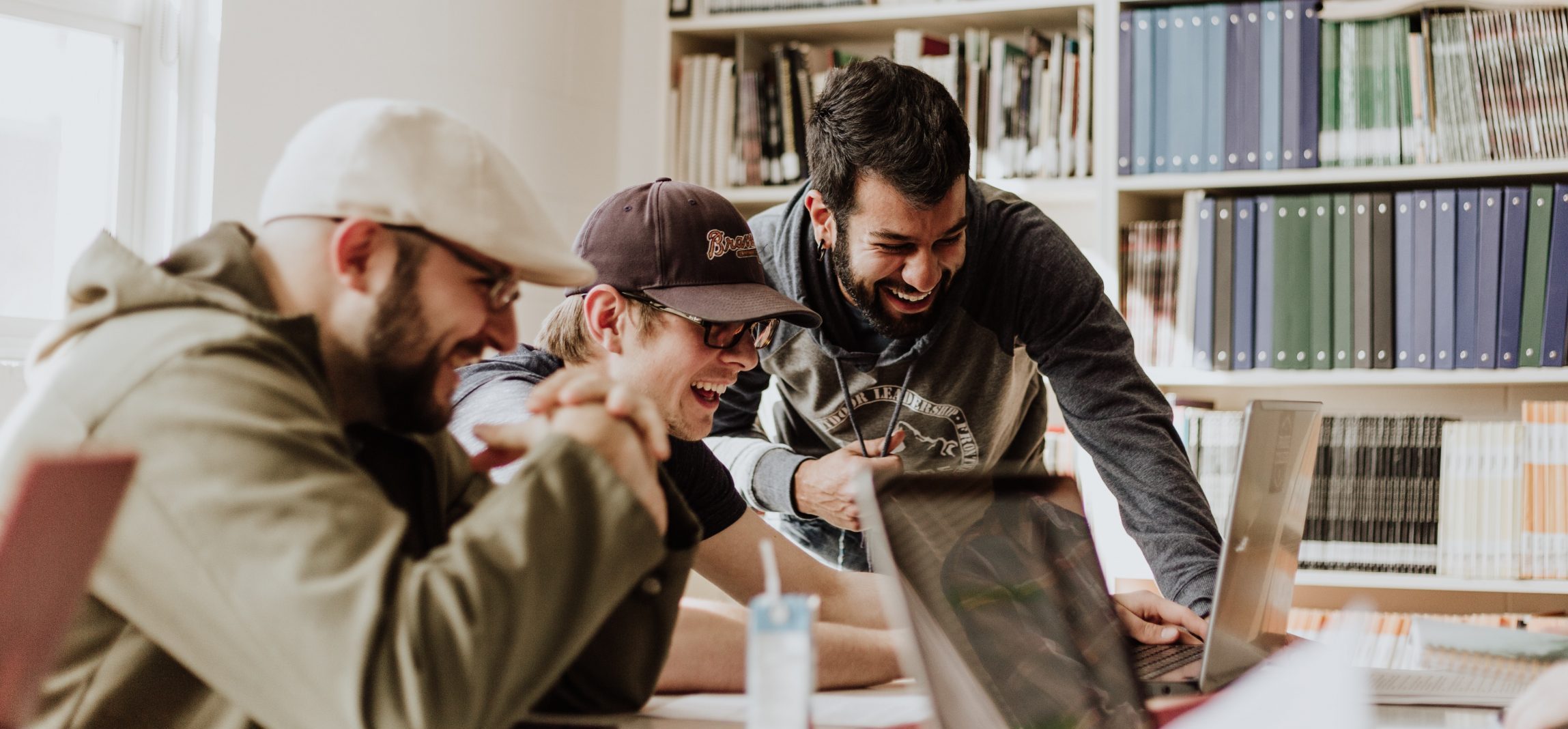 Drie personen kijken lachend naar een laptop op tafel, op de achtergrond staat een boekenkast, ze volgen een compliance training.